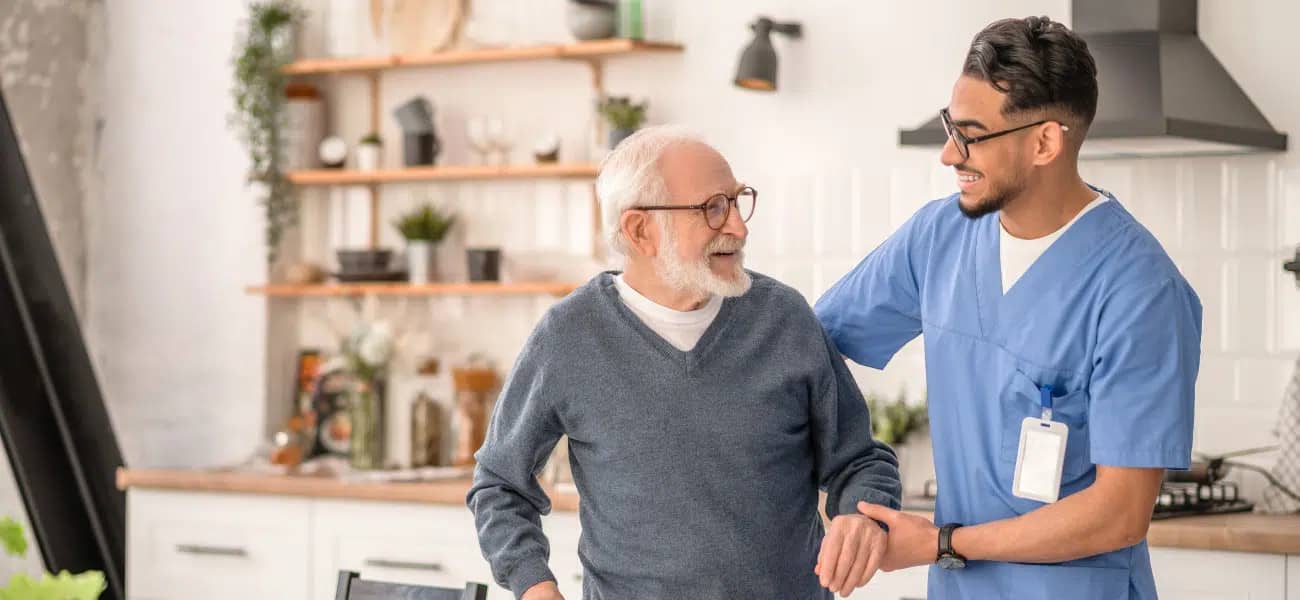 A caregiver steadying an older man in a bright kitchen